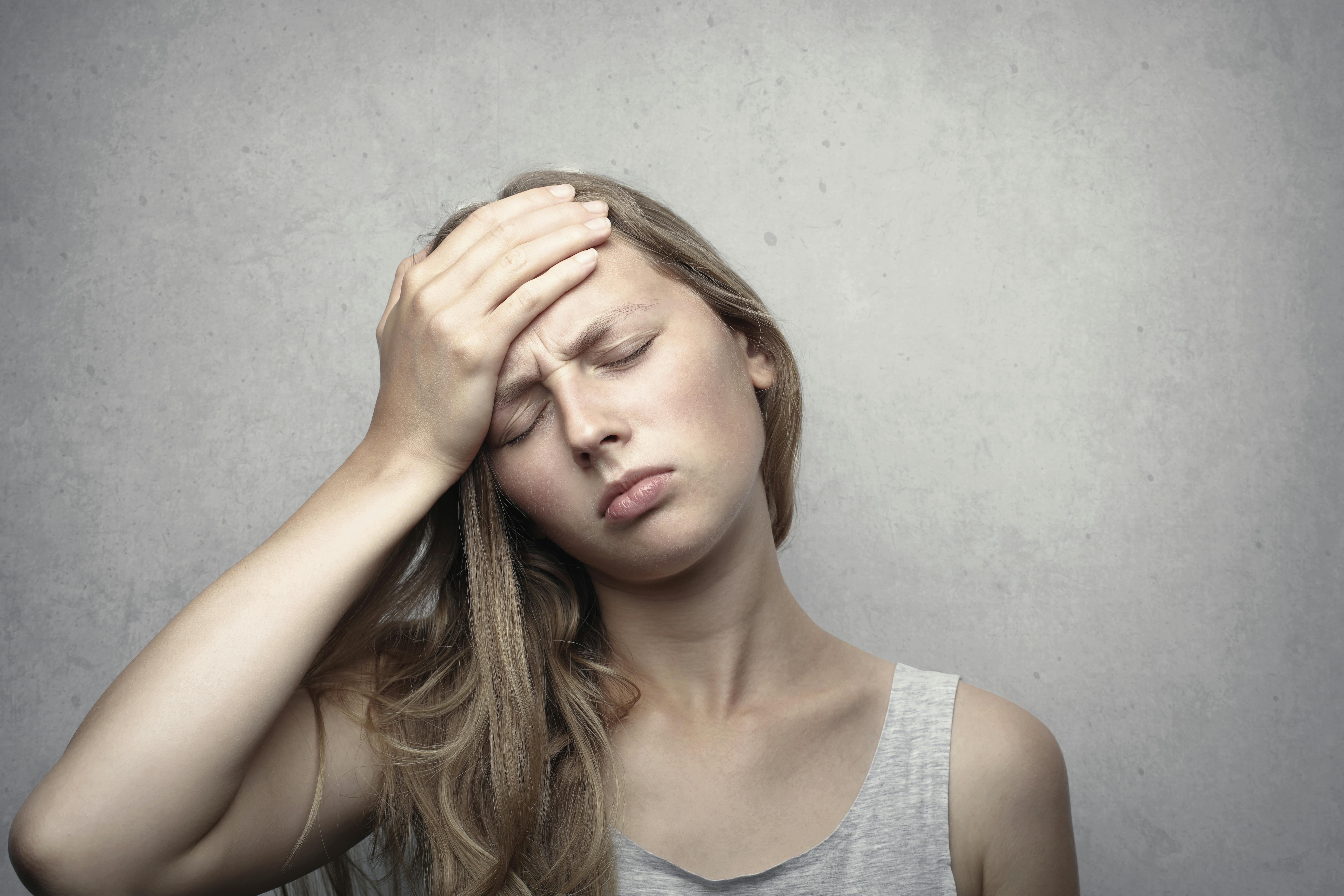 A woman holding her forehead with a pained expression, eyes closed, wearing a sleeveless top, standing against a neutral background. She appears to be experiencing fatigue or a headache.