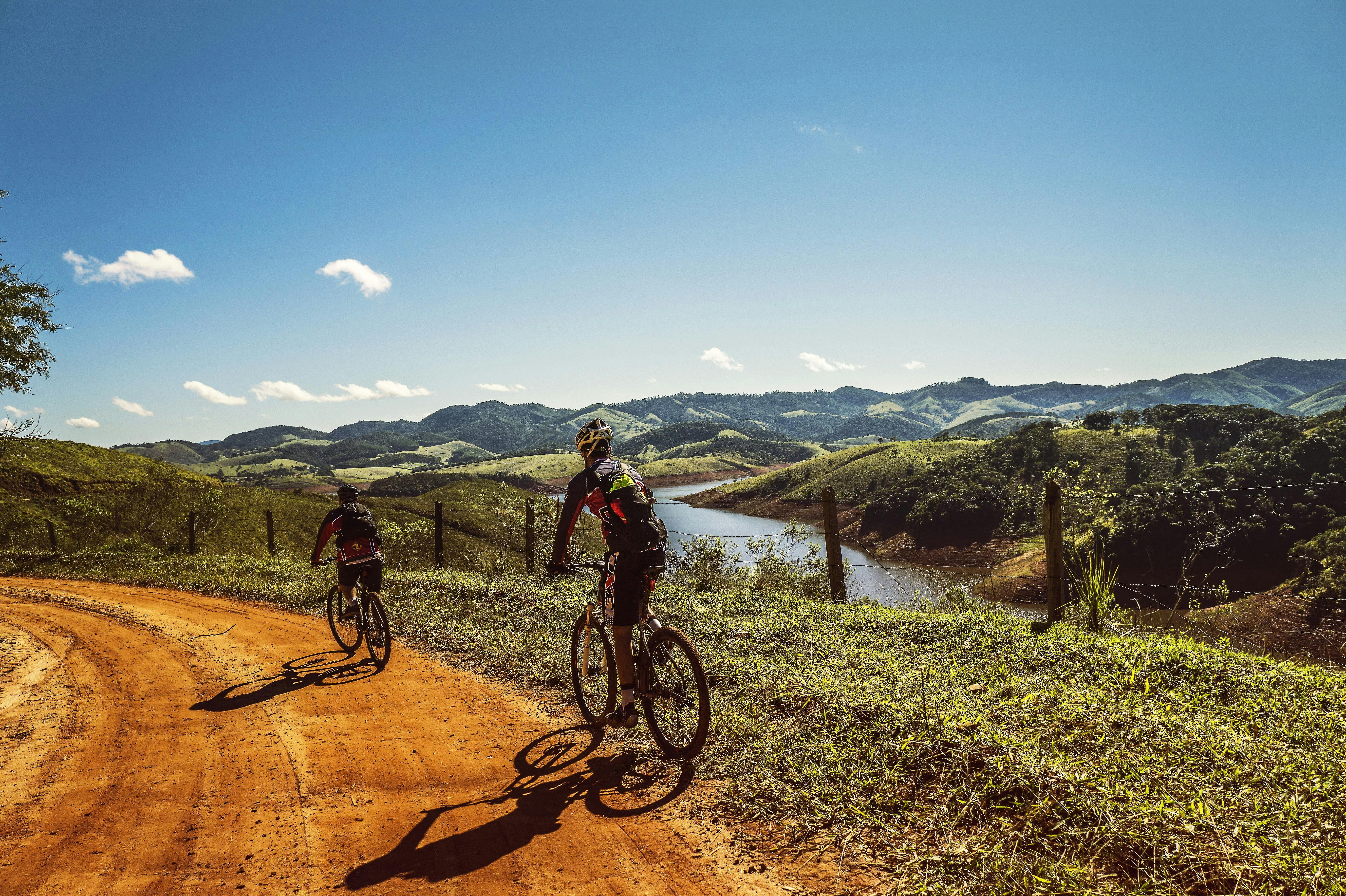 Two cyclists riding along a winding dirt path in a scenic landscape with rolling green hills and a river under a clear blue sky.