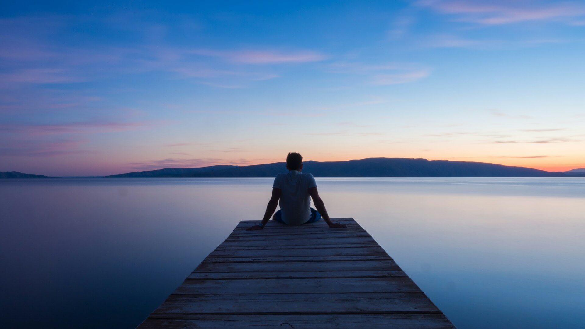 Person sitting alone at the end of a wooden dock overlooking a still lake at sunset, symbolizing the quiet moments before anger strikes.