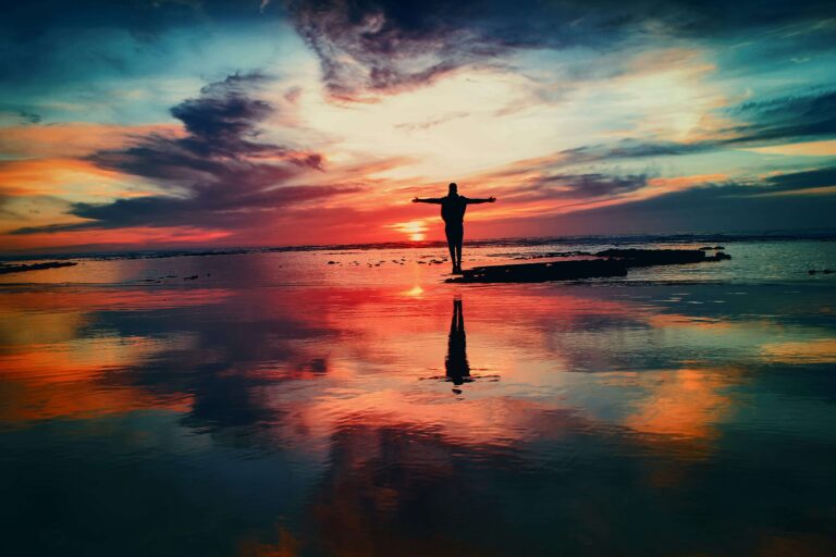 Silhouette of a person standing with open arms at sunset over reflective water, symbolizing peace and the idea that spirituality still matters.