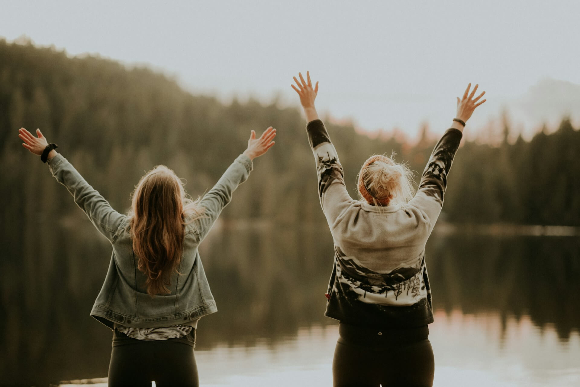 Two people standing by a calm lake with their arms raised toward the sky, symbolizing connection, renewal, and the idea of spirituality in healing.