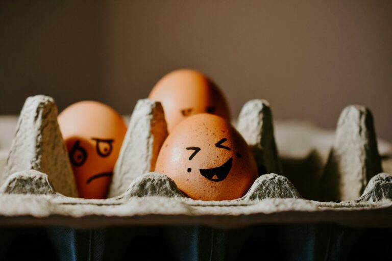 Eggs in a carton with drawn facial expressions, including a cheerful face in front and a sad, suppressed face behind it, symbolizing the idea of suppressing emotions.