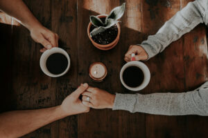 Two people holding hands across a wooden table with coffee cups and a candle, symbolizing warmth, support, and the lifeline of relationships.