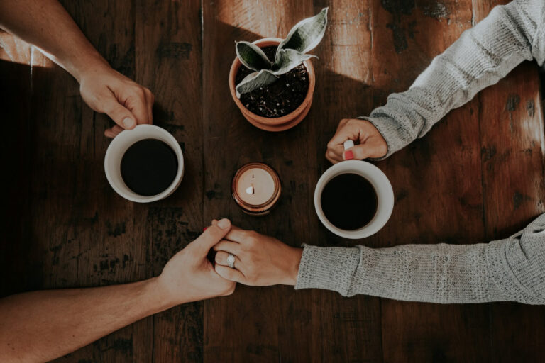Two people holding hands across a wooden table with coffee cups and a candle, symbolizing warmth, support, and the lifeline of relationships.