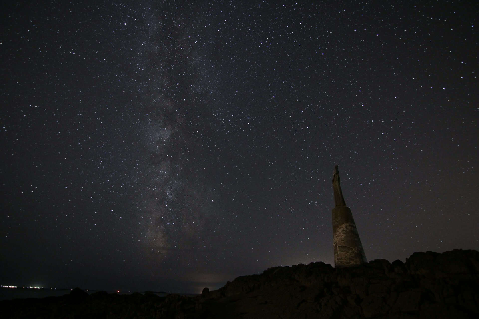 Star-filled night sky with the Milky Way stretching overhead and a distant statue silhouetted on rocky ground, symbolizing the mystery behind horoscopes and cosmic guidance.